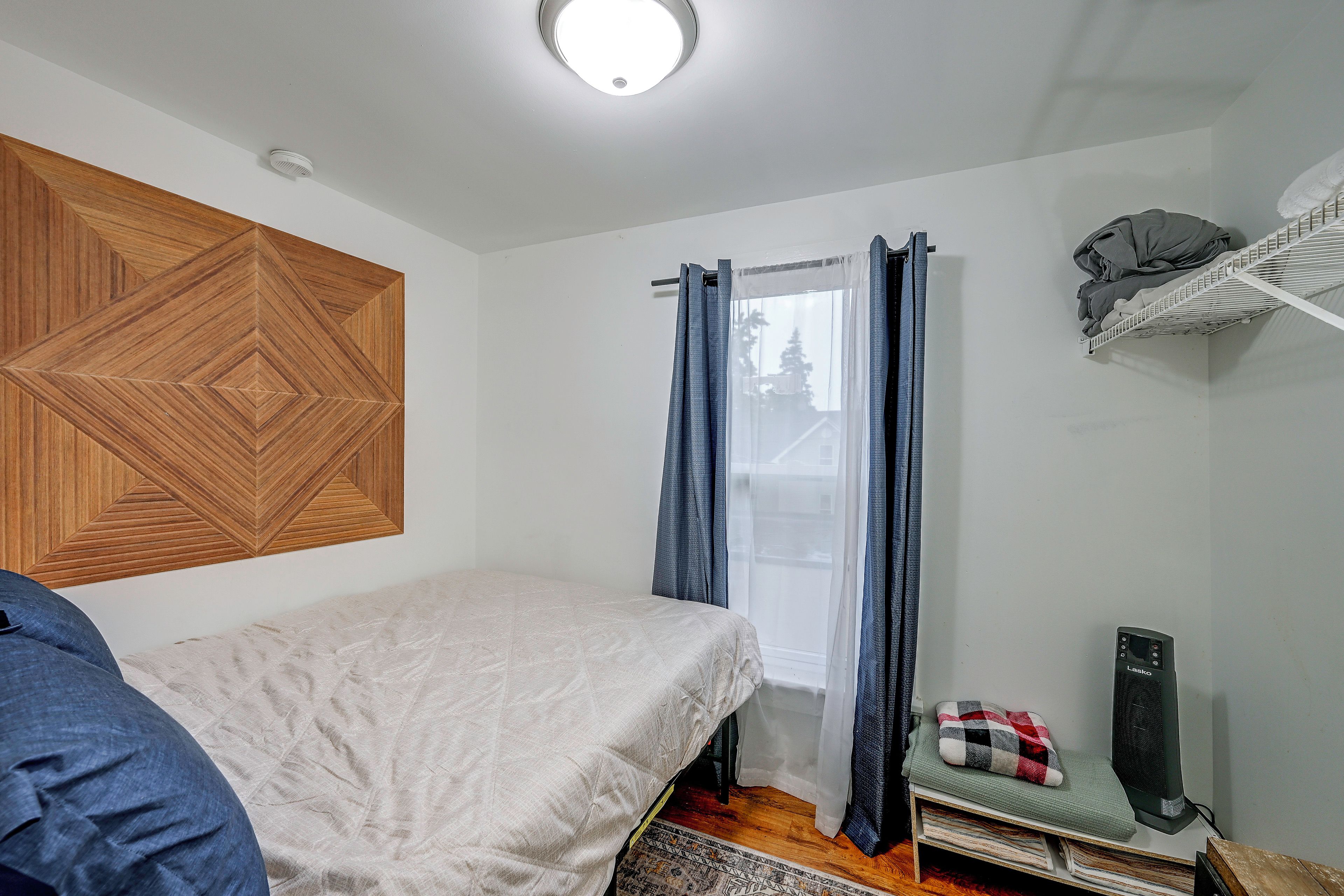 Un dormitorio con una cama, un panel de pared de madera, una ventana con cortinas, un radiador y una estantería con objetos.