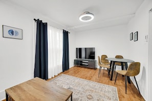 A modern living room with a flat-screen TV, a dining table with chairs, a wooden coffee table, and a patterned rug.
