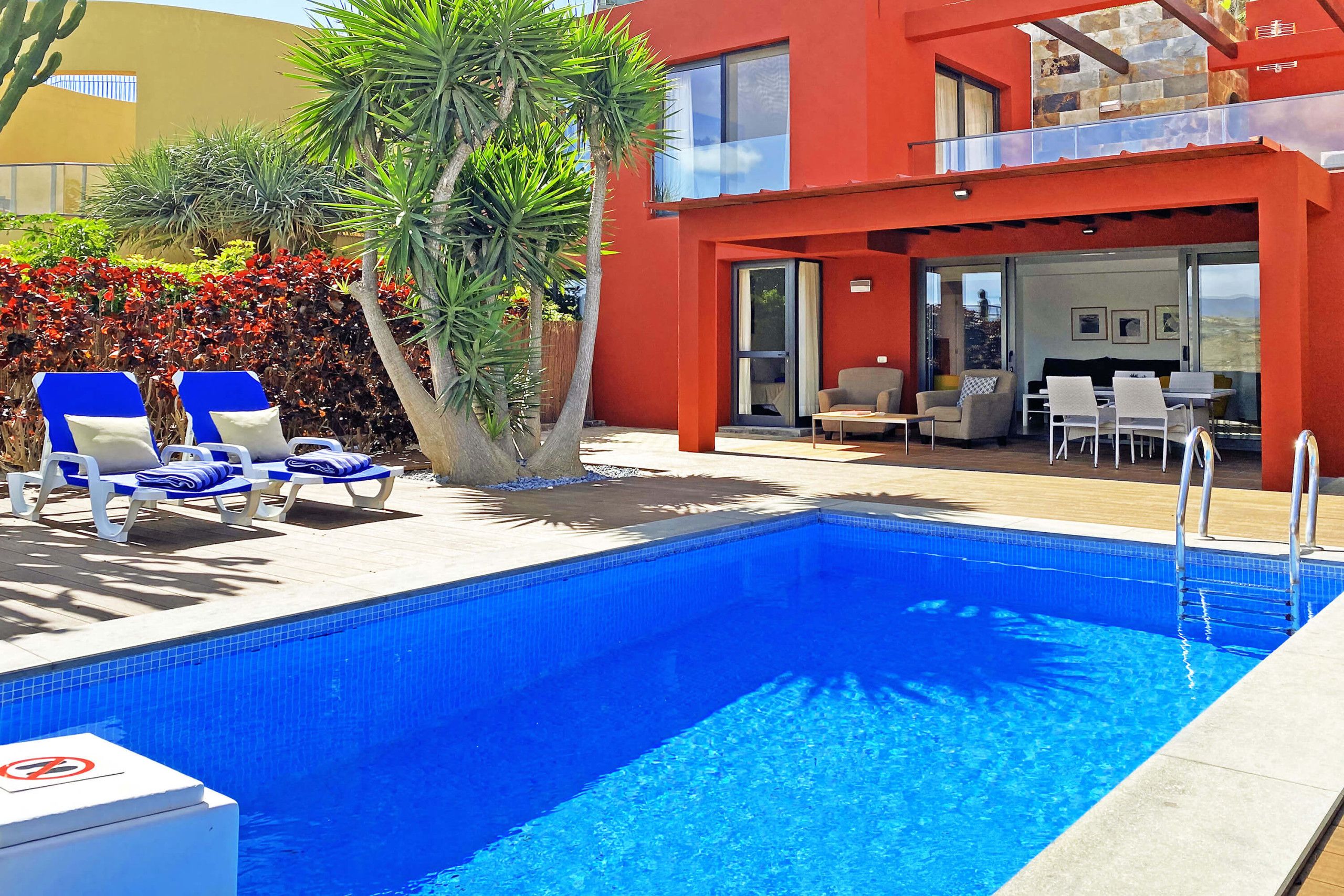 Ein Schwimmbecken mit Liegestühlen, ein Essbereich mit Tischen und Stühlen sowie ein rotes Gebäude mit einem Balkon.