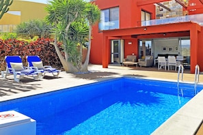 A swimming pool with lounge chairs, a dining area with tables and chairs, and a red building with a balcony. - Beautiful house at Salobre Golf Resort with private pool (San Bartolomé de Tirajana)