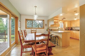 A spacious dining area with a wooden table and chairs, a large window with a view of trees, and a kitchen with wooden cabinets and a bar area. - Riverfront Group Getaway Near Glacier NP (Columbia Falls)