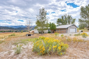 A rural house with a garage, surrounded by dry grassland and yellow flowers, with mountains in the background.