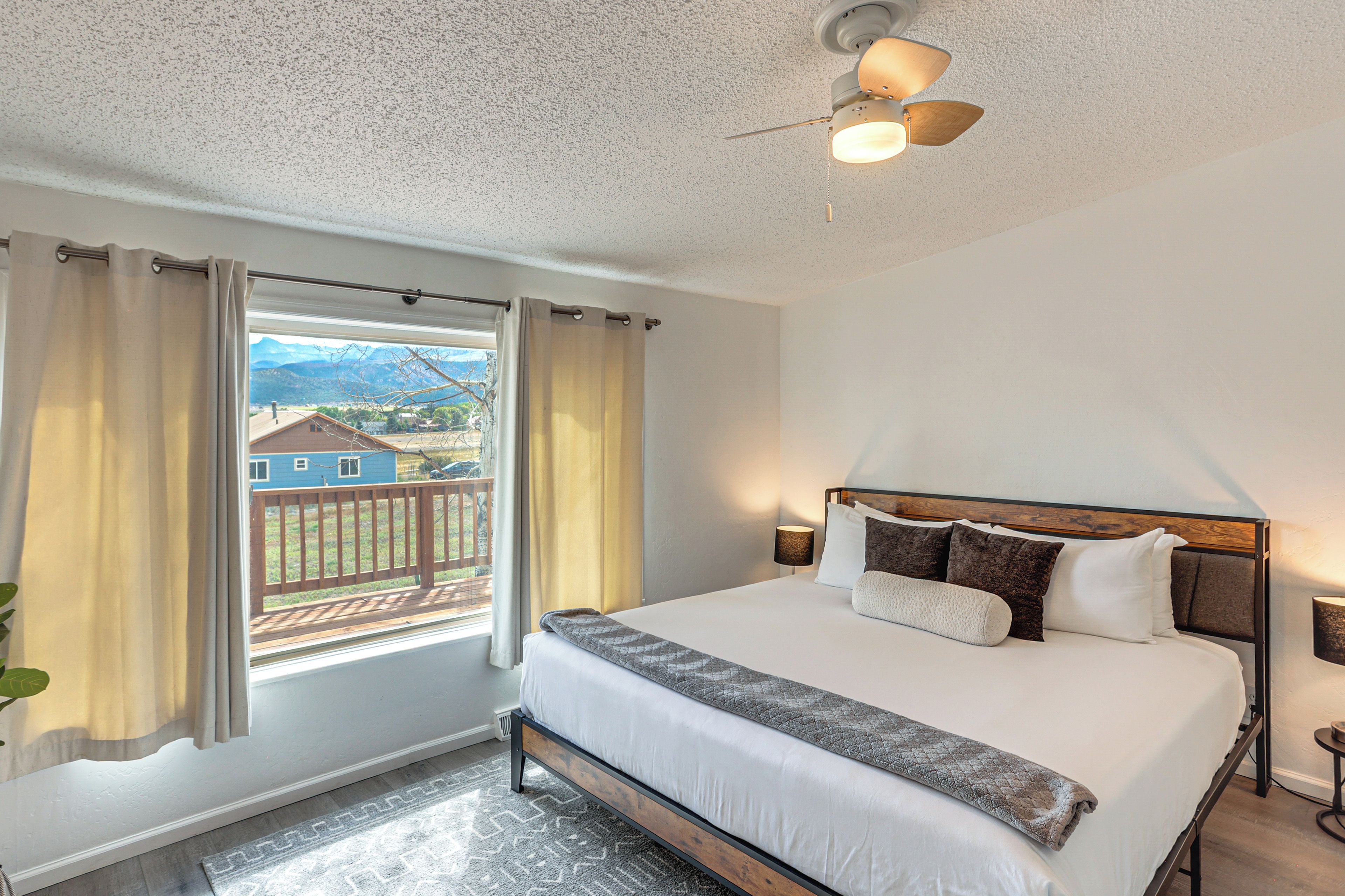 Un dormitorio con cama, ventana con cortinas, ventilador de techo y vistas a una casa y montañas.
