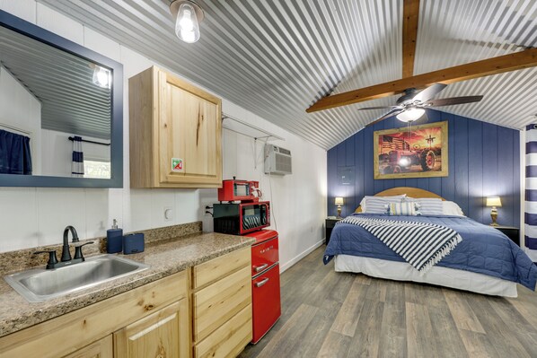 A compact bedroom with a bed, bedside tables, a red appliance, a ceiling fan, and a large window with blinds.