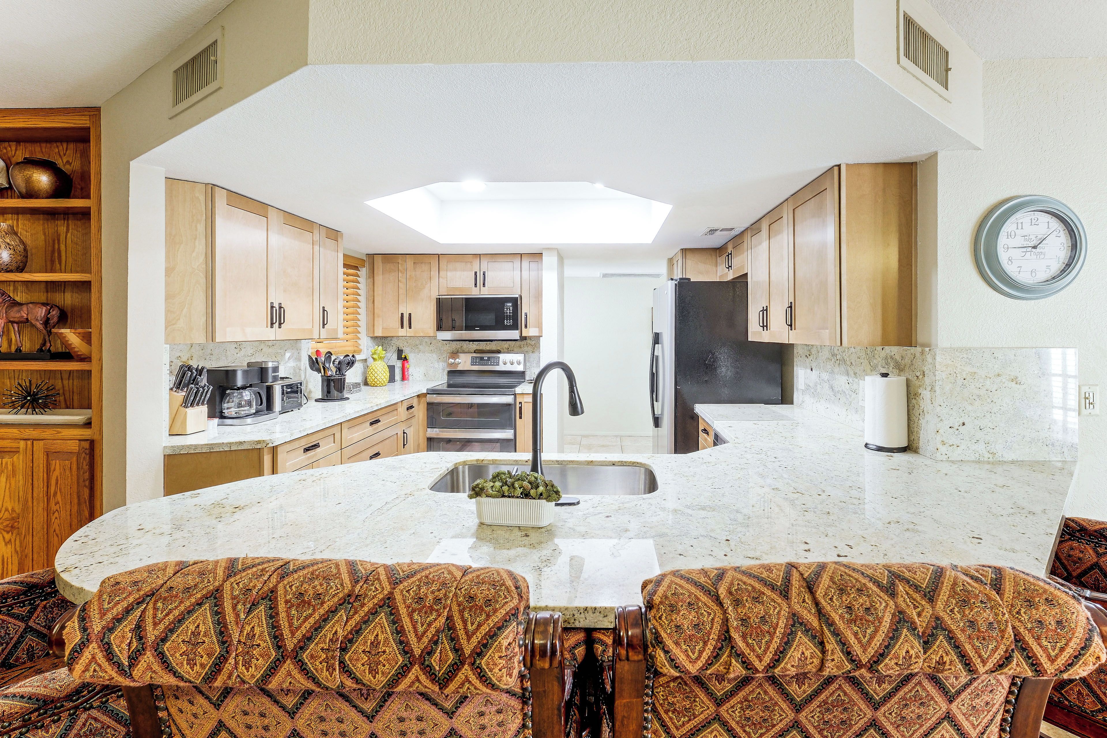 A kitchen with a central island, wooden cabinets, a microwave, oven, and a clock on the wall.