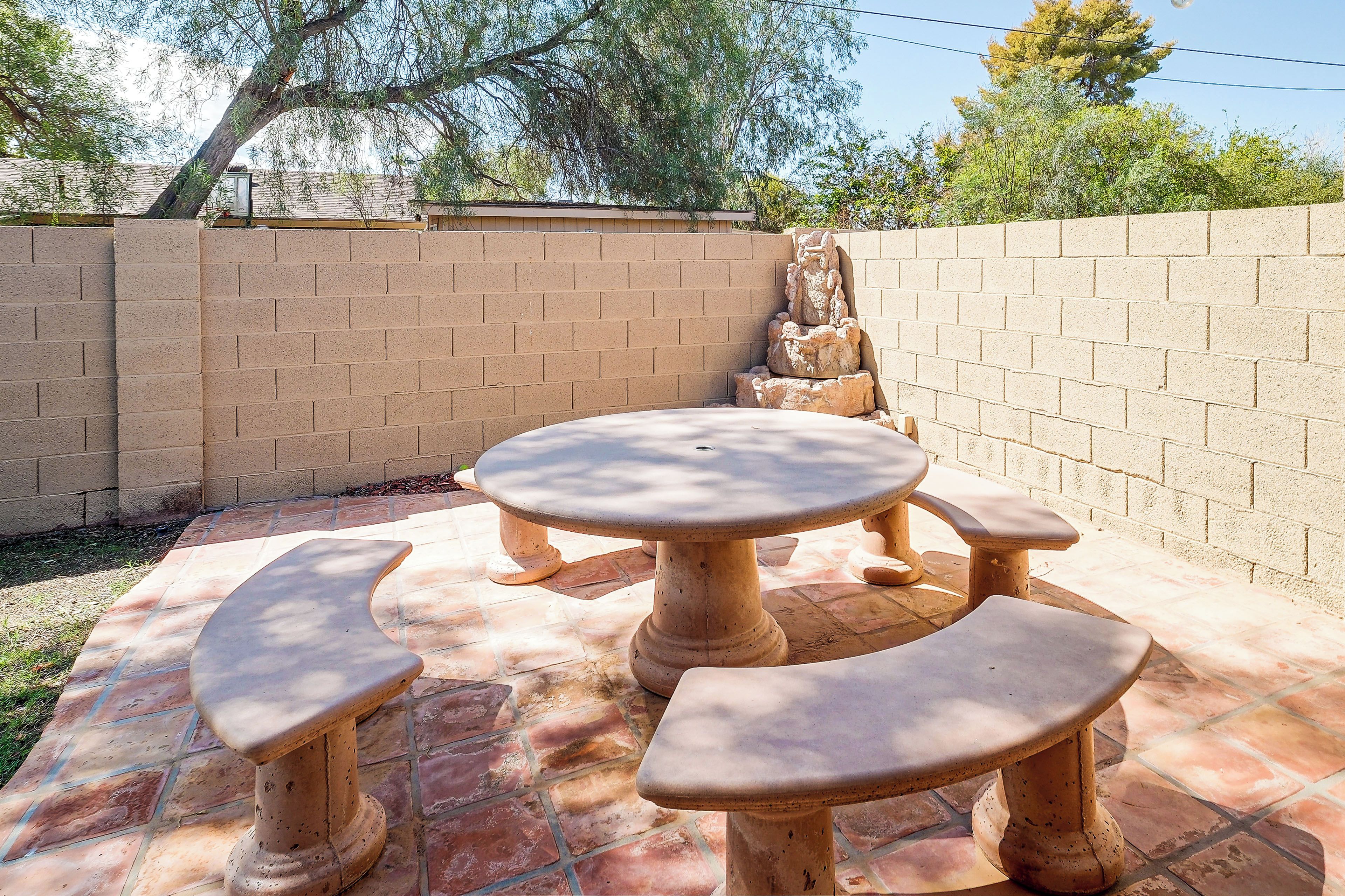 Um pátio externo com mesa redonda e bancos, cercado por um muro de tijolos e um elemento decorativo de pedra.