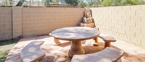 Um pátio externo com mesa redonda e bancos, cercado por um muro de tijolos e um elemento decorativo de pedra.
