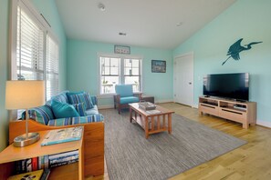 A living room with a sofa, a coffee table, a TV stand, and a bookshelf. - Sunset & Waterway Views! Holden Beach Getaway (Holden Beach)