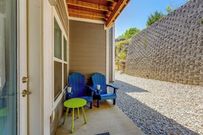 A covered outdoor seating area with two blue chairs and a green table, adjacent to a building with a stone wall. - Condo Near Branson Strip: Close to It All! (Branson)