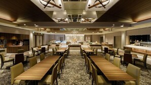 Un espace de restauration moderne dans un hôtel, avec de nombreuses tables et chaises, un lustre central et une cuisine visible au fond.