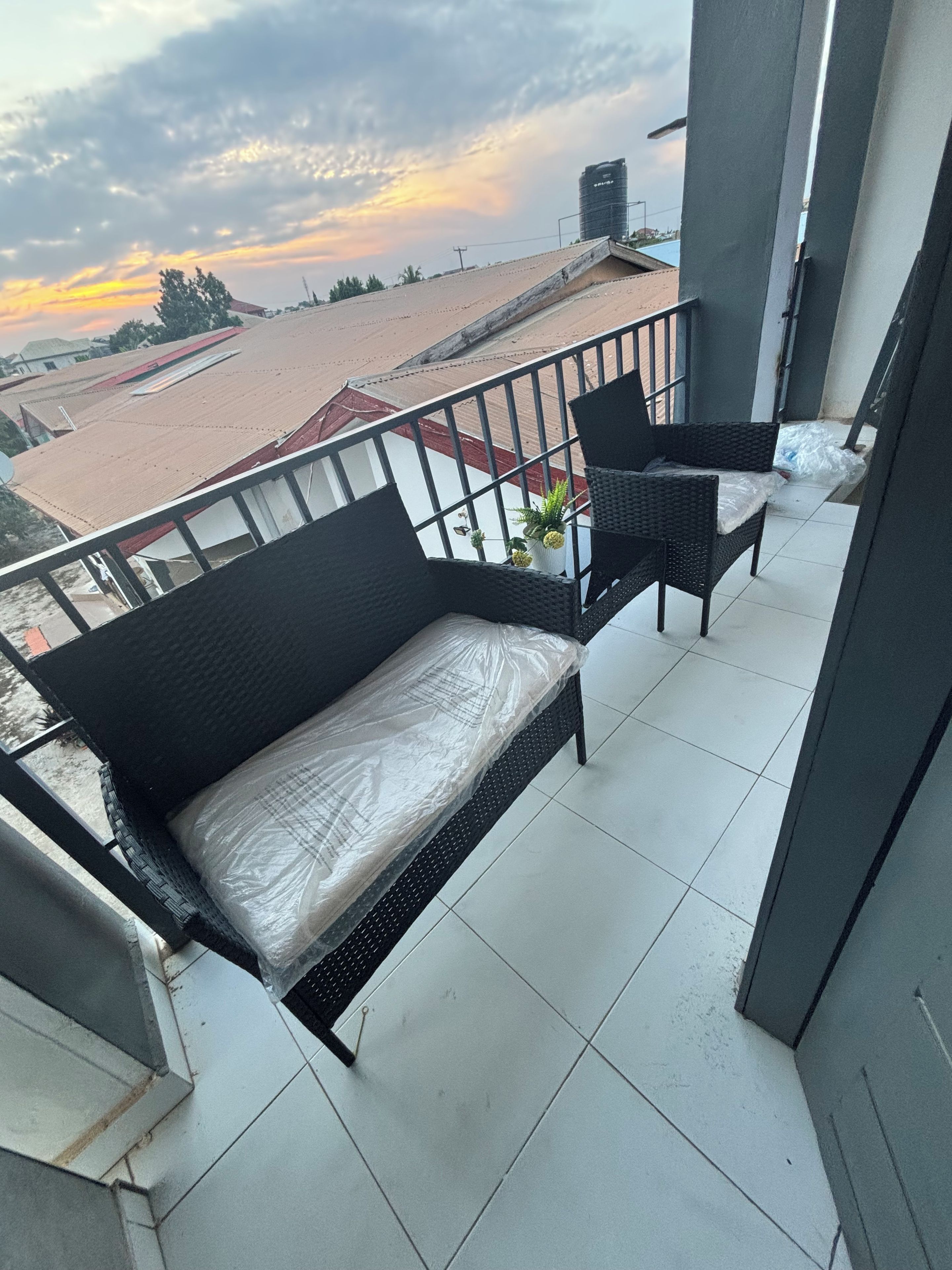 Un balcon avec un canapé noir et deux fauteuils, un sol carrelé et une vue sur des immeubles et un coucher de soleil.