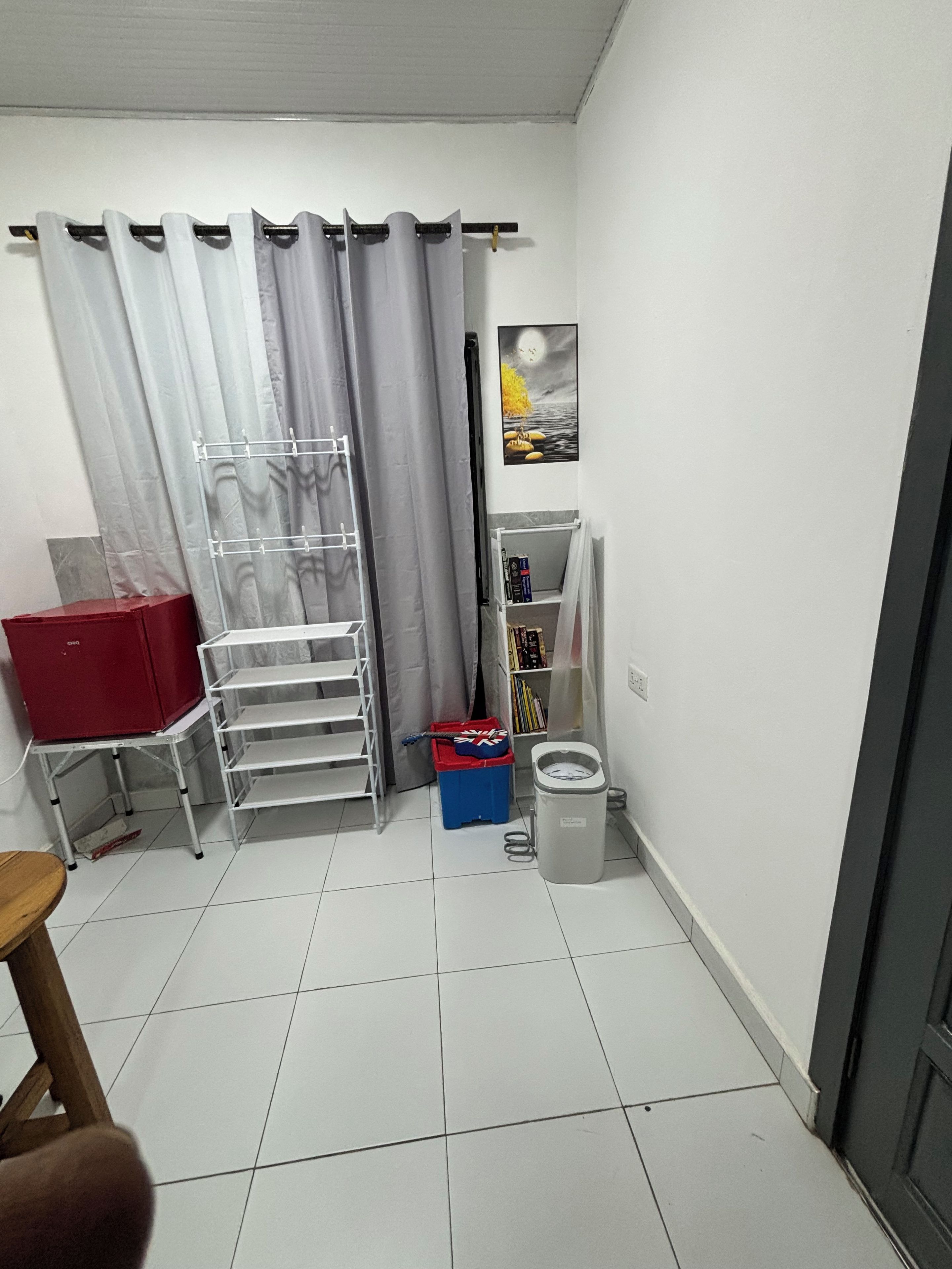 Une petite pièce propre, avec un sol carrelé de carreaux blancs, une armoire rouge, un étendoir métallique, une boîte de rangement bleue, une poubelle blanche et une bibliothèque.