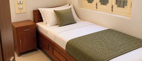 Une chambre avec un lit simple, un cadre en bois, une table de chevet, une petite table d’appoint, une fenêtre avec du verre dépoli et un interrupteur fixé au mur.