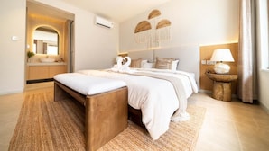 Quarto moderno com uma cama grande, um banco de madeira, uma mesa de madeira redonda com uma luminária e um espelho.