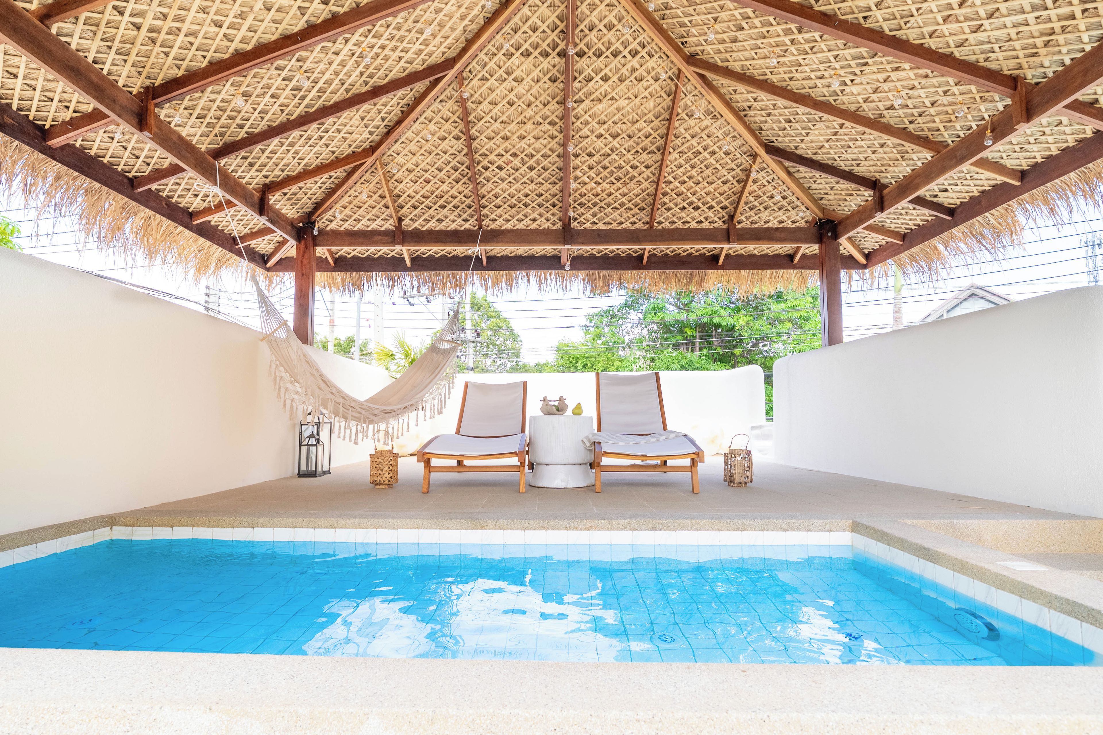 A pool area with a thatched roof, two lounge chairs, and a small table.