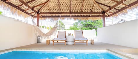 A pool area with a thatched roof, two lounge chairs, and a small table.