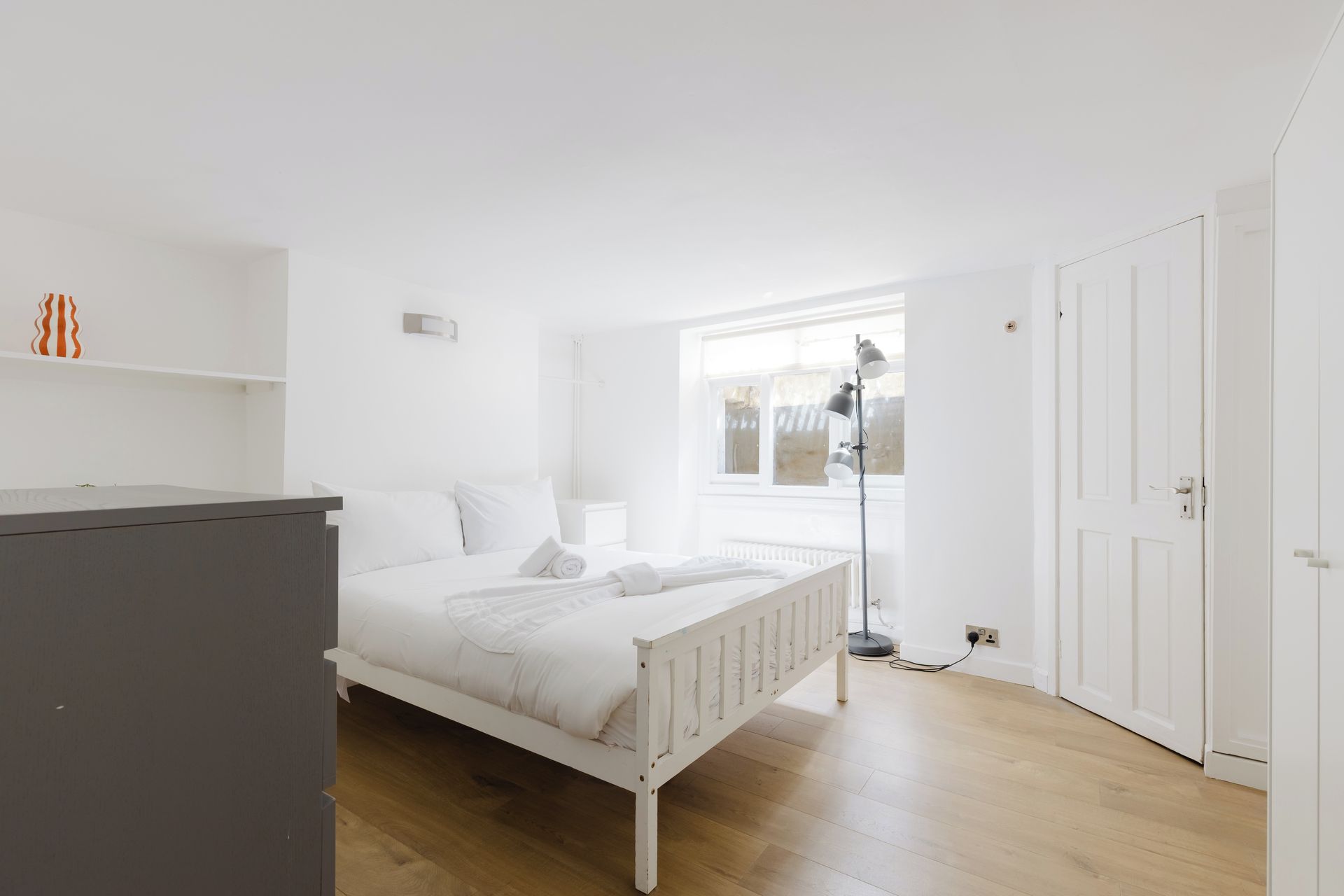 Een slaapkamer met een wit bed, een nachtkastje, een vloerlamp en een kledingkast.