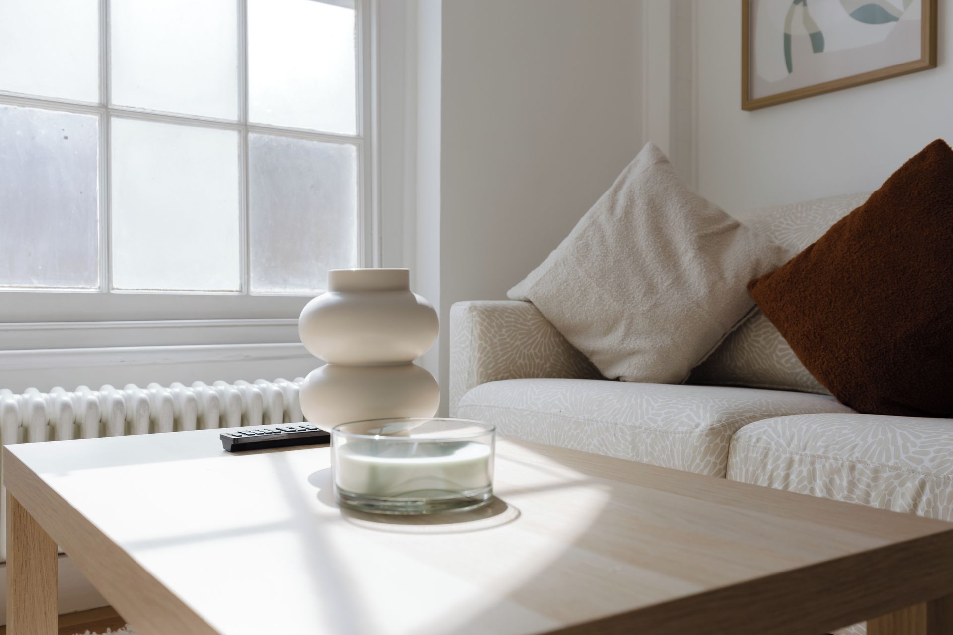 Una habitación con un sofá blanco, un cojín marrón, un jarrón blanco, un control remoto y un candelabro de vidrio sobre una mesa de madera.