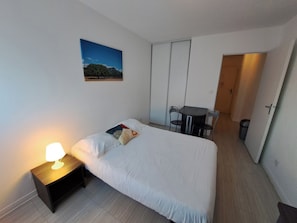 Un dormitori amb un llit, una taula de nit amb una làmpada, un petit escriptori amb dues cadires, un quadre a la paret i una porta que condueix a una altra habitació.