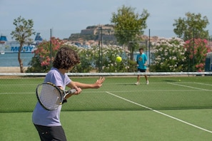 A tennis court with a net in the middle, surrounded by greenery and a body of water in the background. - Club Airone (Portoferraio)