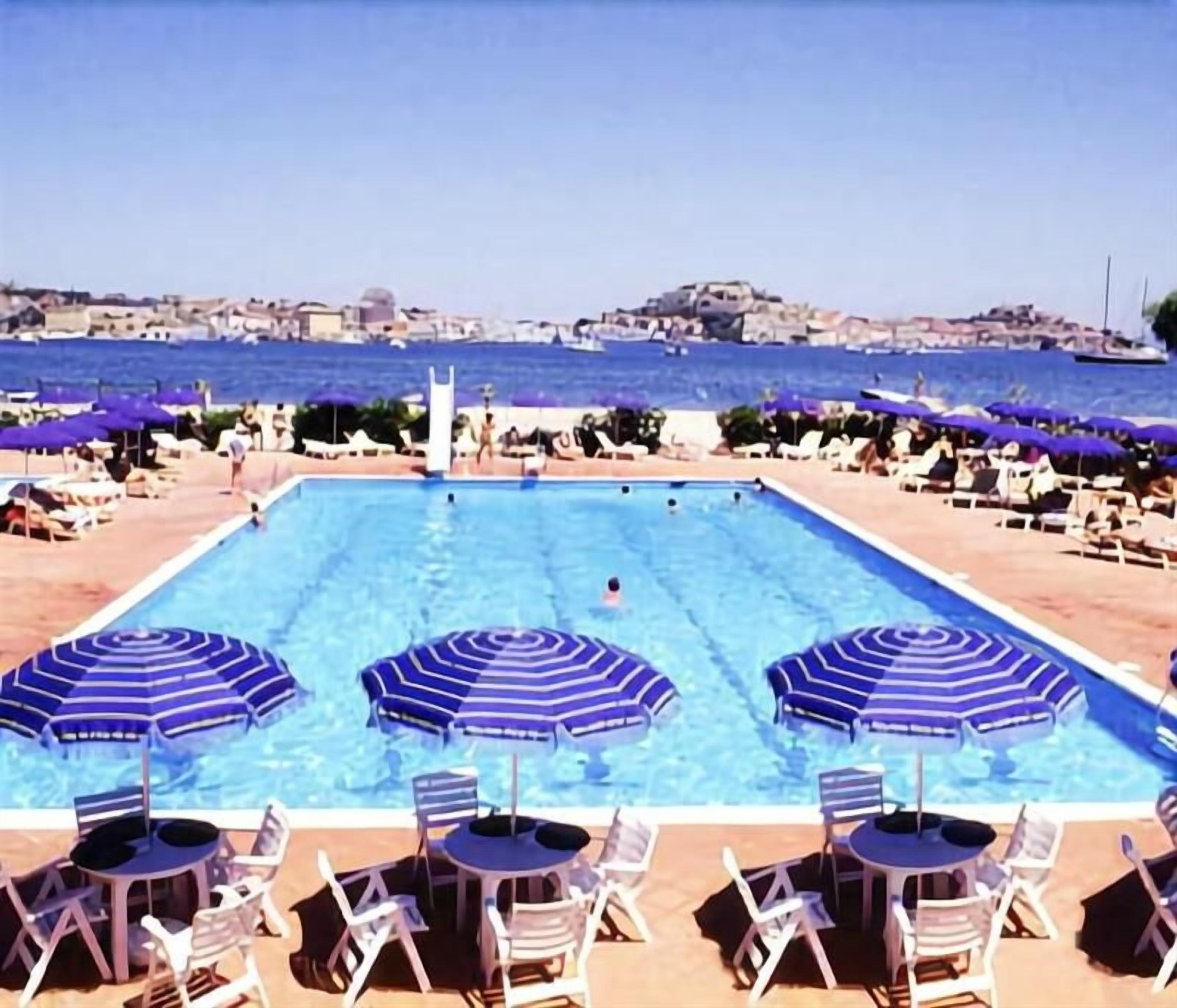 A swimming pool with lounge chairs and umbrellas, a view of the sea, and a distant cityscape.