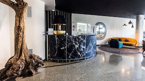 A modern lobby with a curved marble reception desk, a large wooden sculpture, a black lamp, and a yellow sofa. - Mefuta Hotel (Gardone Riviera)