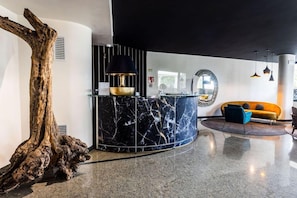 Eine moderne Lobby mit einem geschwungenen Marmor-Empfangstresen, einer großen Holzskulptur, einer schwarzen Lampe und einem gelben Sofa.