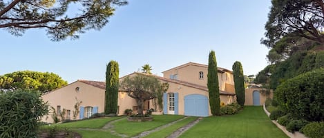 Una villa de estilo mediterráneo con un camino de piedra que lleva a una puerta azul, rodeada de jardines cuidados y altos cipreses.