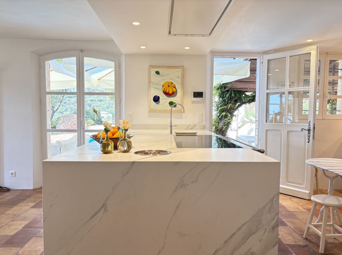 A modern kitchen with a marble island, large windows, and a view of greenery outside.