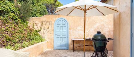 Une terrasse avec un barbecue, une table et un parasol.