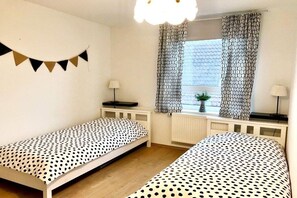 Habitació amb dos llits individuals, una finestra amb cortines estampades, un radiador i una làmpada penjada a la paret.