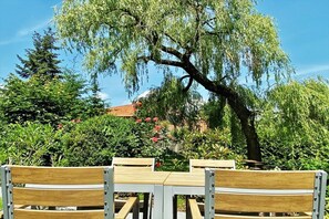 Un espacio exterior con bancos de madera y una mesa, rodeado de abundante vegetación y un gran árbol.