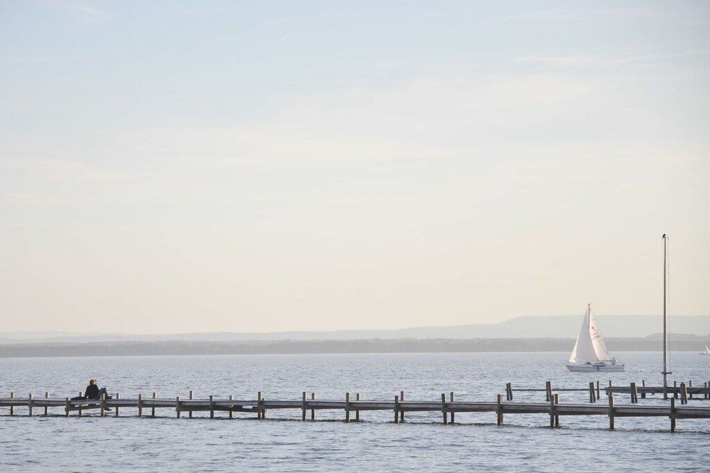 Ruhiges Ufer mit einer hölzernen Pier, die ins Wasser führt, ein Segelboot in der Ferne und eine Person, die auf der Pier sitzt.
