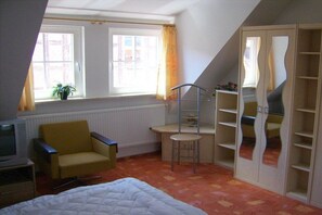 Ein Zimmer mit einem Bett, einem Sessel, einem Schreibtisch, einem Fernseher, einem Fenster mit Vorhängen und einem Kleiderschrank mit Spiegel.