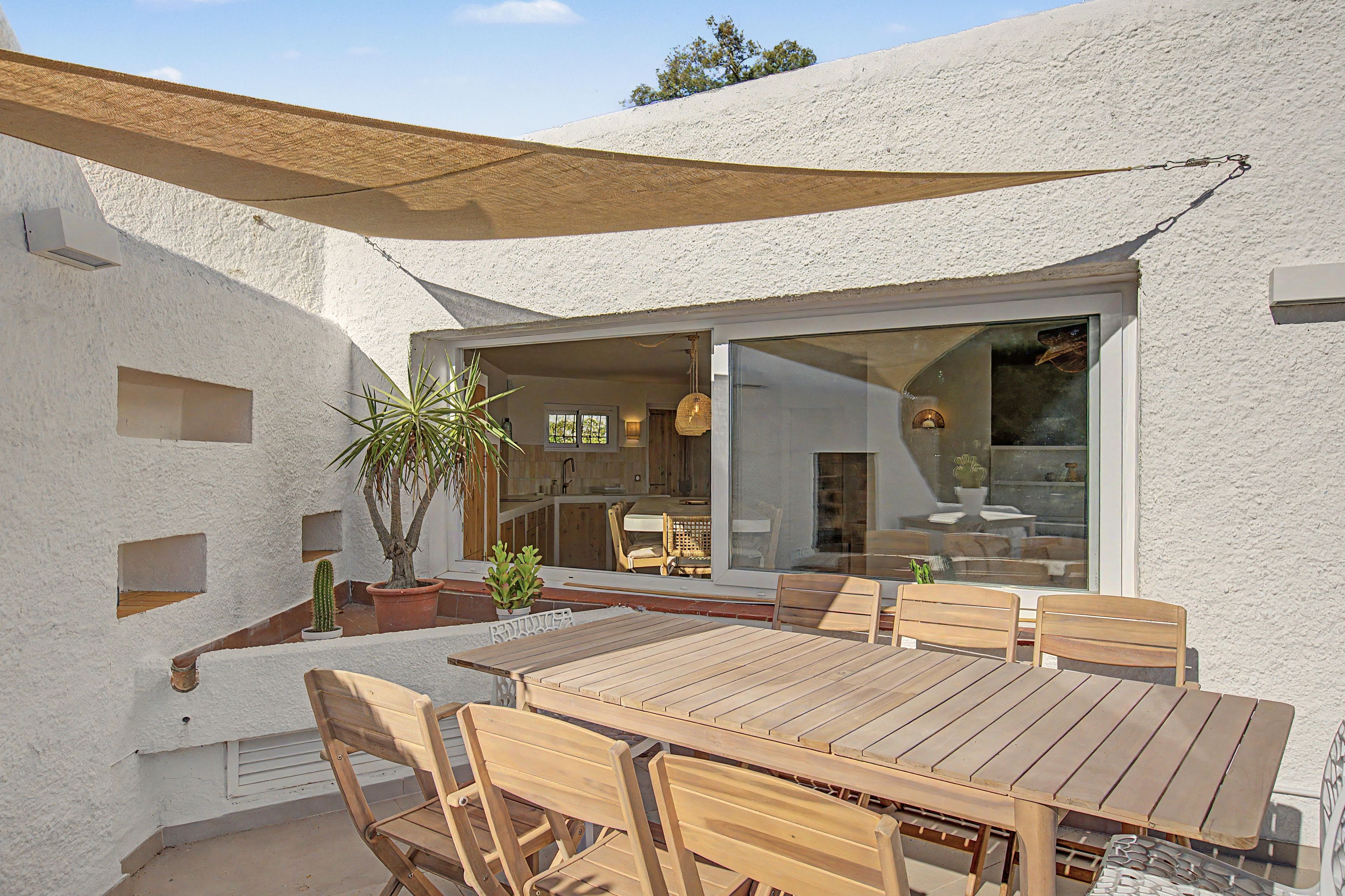 An outdoor patio with a wooden table and chairs, surrounded by white walls and a shaded area.