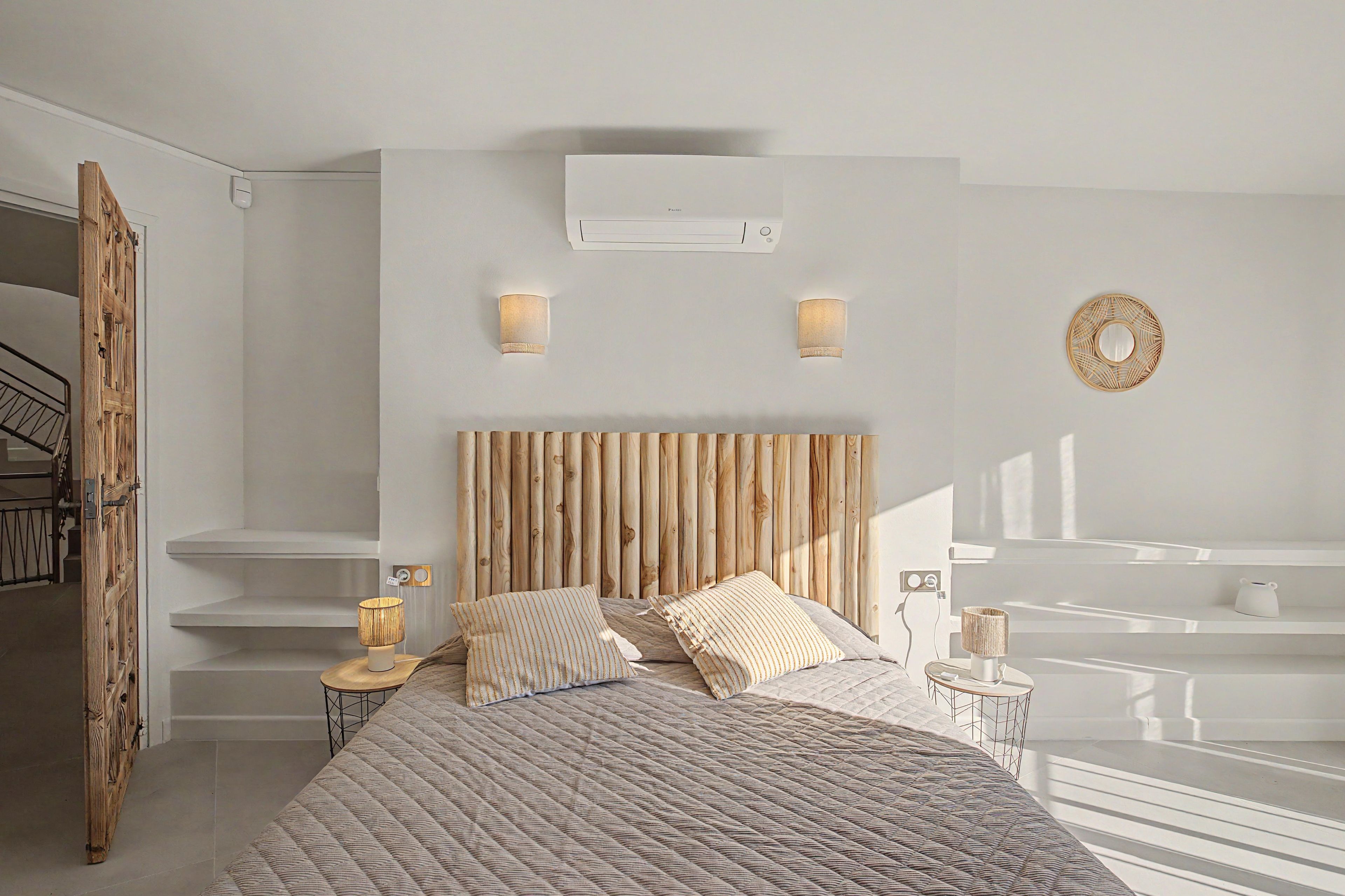 A bedroom with a wooden headboard, two bedside lamps, a bed with patterned bedding, and a wall-mounted air conditioner.