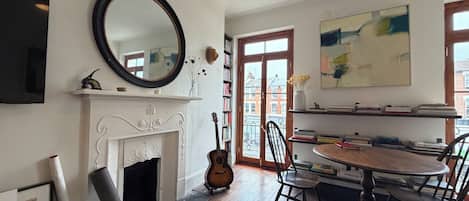 Una habitación con chimenea, una mesa de comedor con sillas, una guitarra, una estantería y un cuadro abstracto grande.