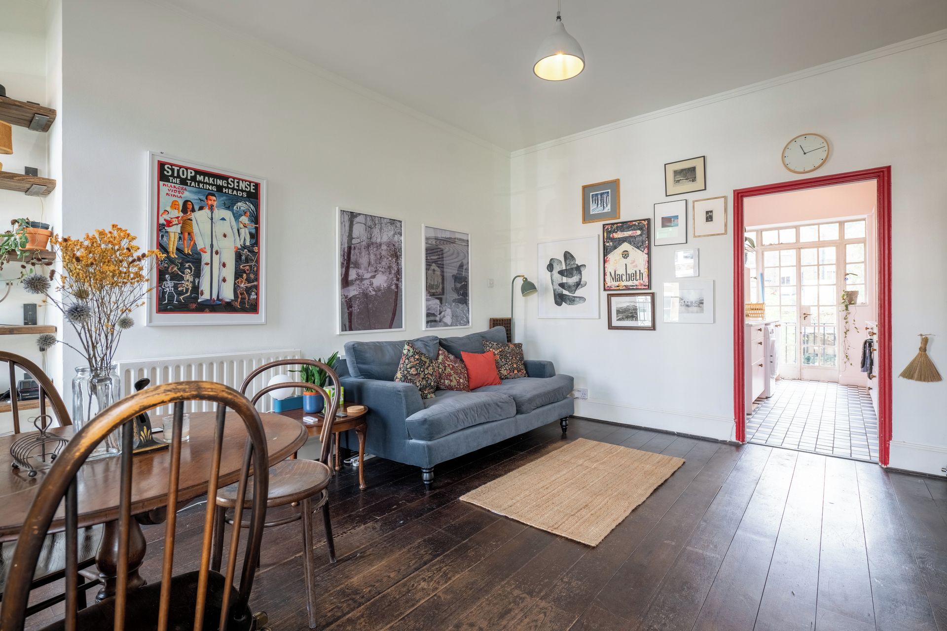 Ein Wohnzimmer mit einem blauen Sofa, einem Holzesstisch und Stühlen. An den Wänden befinden sich gerahmte Bilder und eine rote Tür.