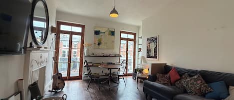 Un salón amplio con un sofá oscuro, una mesa de comedor y una cocina visible a través de una puerta abierta.