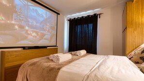 A hotel room with a bed, a projector screen displaying a scenic image, a wooden headboard, and a window with curtains. - Fiemmea – Hidden Chalet, Valley View in Cavalese (Cavalese)