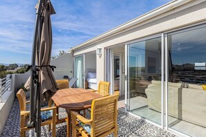 Ein Balkon mit Holztisch und -stühlen, einem schattenspendenden Sonnenschirm und einer Glastür zum Schlafzimmer.