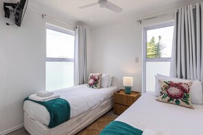 Una habitación de hotel con dos camas, un ventilador de techo, una mesita de noche con lámpara y una ventana con cortinas.