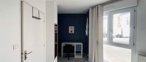 Un pasillo con una puerta a la izquierda, una ventana a la derecha y una pequeña mesa con una silla en el centro.