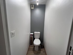 A narrow bathroom with a toilet, a small trash bin, and a vent on the wall.