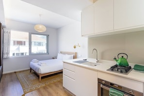 A compact hotel room with a bed, a small kitchenette, a window with blinds, and a view of buildings outside. - Kloof Street Studio Table Mountain views by ctha (Cape Town)
