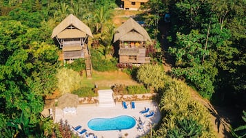 An aerial view of a resort with a swimming pool, lounge chairs, and thatched-roof buildings surrounded by dense vegetation.