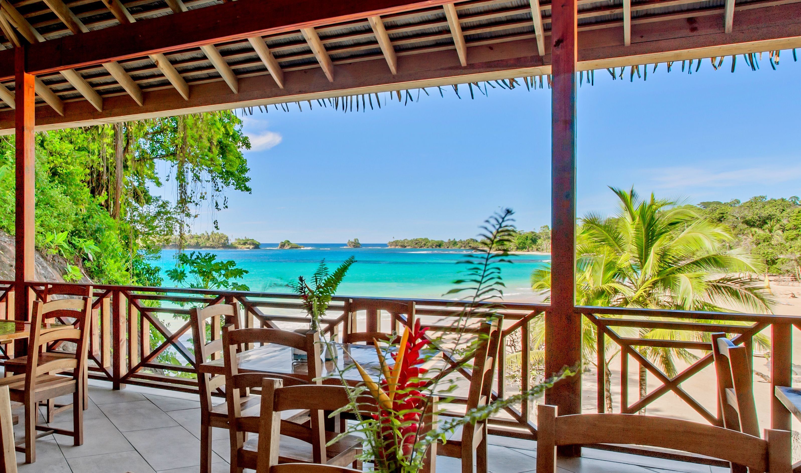 A covered outdoor seating area with wooden chairs and tables, overlooking a beach and turquoise waters.