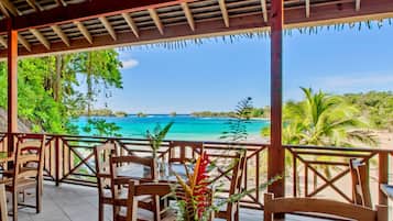 A covered outdoor seating area with wooden chairs and tables, overlooking a beach and turquoise waters.