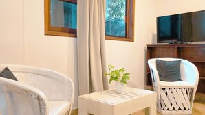 A room with two white wicker chairs, a small white table with a plant, a television on a wooden stand, and a window with curtains. - Garibaldi Inn at the Bay (Mahahual)