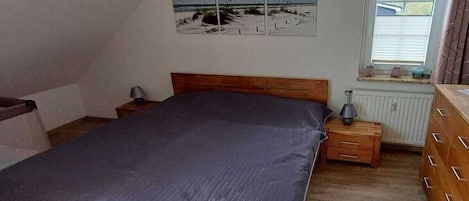 A bedroom with a bed, a nightstand, a dresser, a window with blinds, and a beach-themed wall decoration.